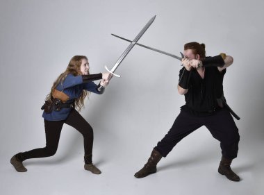 Kızıl saçlı bir çiftin tam boy portresi, ortaçağ Viking 'i giymiş bir adam ve kadın ilham verici fantezi kostümleri, duruş pozisyonu kılıç tutuş, beyaz stüdyo arka planında izole edilmiş.