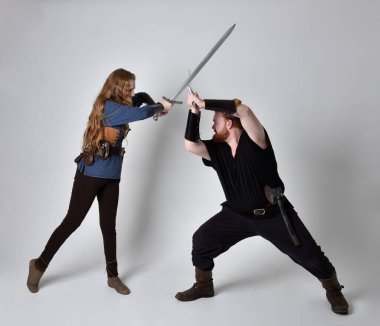 Kızıl saçlı bir çiftin tam boy portresi, ortaçağ Viking 'i giymiş bir adam ve kadın ilham verici fantezi kostümleri, duruş pozisyonu kılıç tutuş, beyaz stüdyo arka planında izole edilmiş.