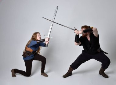 Kızıl saçlı bir çiftin tam boy portresi, ortaçağ Viking 'i giymiş bir adam ve kadın ilham verici fantezi kostümleri, duruş pozisyonu kılıç tutuş, beyaz stüdyo arka planında izole edilmiş.