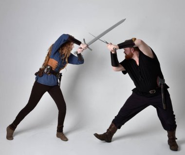 Kızıl saçlı bir çiftin tam boy portresi, ortaçağ Viking 'i giymiş bir adam ve kadın ilham verici fantezi kostümleri, duruş pozisyonu kılıç tutuş, beyaz stüdyo arka planında izole edilmiş.