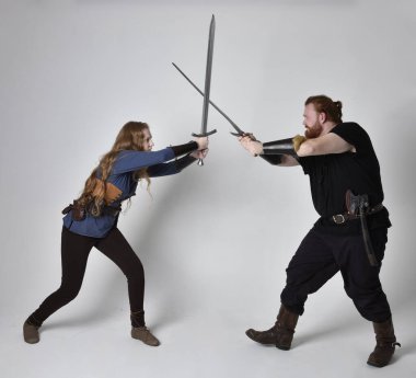Kızıl saçlı bir çiftin tam boy portresi, ortaçağ Viking 'i giymiş bir adam ve kadın ilham verici fantezi kostümleri, duruş pozisyonu kılıç tutuş, beyaz stüdyo arka planında izole edilmiş.