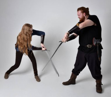 Kızıl saçlı bir çiftin tam boy portresi, ortaçağ Viking 'i giymiş bir adam ve kadın ilham verici fantezi kostümleri, duruş pozisyonu kılıç tutuş, beyaz stüdyo arka planında izole edilmiş.