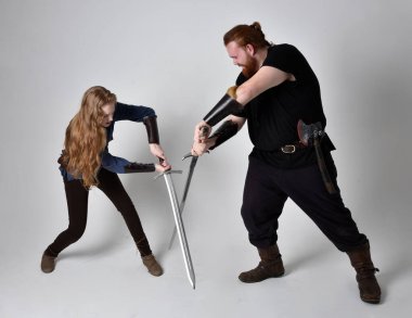 Kızıl saçlı bir çiftin tam boy portresi, ortaçağ Viking 'i giymiş bir adam ve kadın ilham verici fantezi kostümleri, duruş pozisyonu kılıç tutuş, beyaz stüdyo arka planında izole edilmiş.