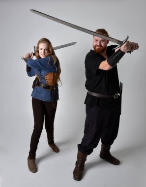 Kızıl saçlı bir çiftin tam boy portresi, ortaçağ Viking 'i giymiş bir adam ve kadın ilham verici fantezi kostümleri, duruş pozisyonu kılıç tutuş, beyaz stüdyo arka planında izole edilmiş.