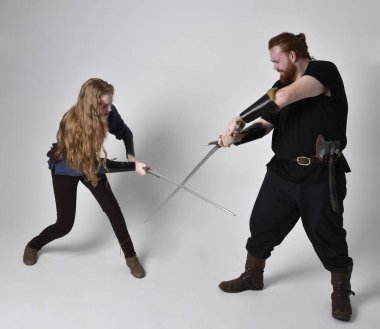 Kızıl saçlı bir çiftin tam boy portresi, ortaçağ Viking 'i giymiş bir adam ve kadın ilham verici fantezi kostümleri, duruş pozisyonu kılıç tutuş, beyaz stüdyo arka planında izole edilmiş.