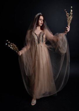 Peri tanrıçası kostümü gibi kremalı bir fantezi elbisesi giyen kızıl saçlı kızın tam boy portresi. Stüdyonun arka planında izole edilmiş zarif hareketli duruş..