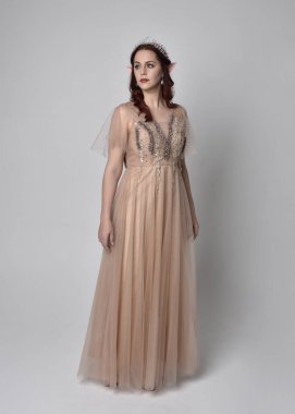 Peri tanrıçası kostümü gibi kremalı fantezi elbisesi ve kristal taç giyen kızıl saçlı bir kızın tam boy portresi. Hafif stüdyo arka planında izole edilmiş zarif hareketli duruş..