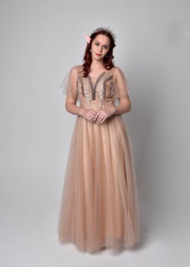 Peri tanrıçası kostümü gibi kremalı fantezi elbisesi ve kristal taç giyen kızıl saçlı bir kızın tam boy portresi. Hafif stüdyo arka planında izole edilmiş zarif hareketli duruş..