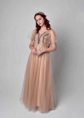 Peri tanrıçası kostümü gibi kremalı fantezi elbisesi ve kristal taç giyen kızıl saçlı bir kızın tam boy portresi. Hafif stüdyo arka planında izole edilmiş zarif hareketli duruş..