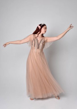 Peri tanrıçası kostümü gibi kremalı fantezi elbisesi ve kristal taç giyen kızıl saçlı bir kızın tam boy portresi. Hafif stüdyo arka planında izole edilmiş zarif hareketli duruş..