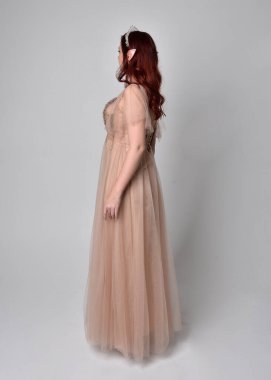 Peri tanrıçası kostümü gibi kremalı fantezi elbisesi ve kristal taç giyen kızıl saçlı bir kızın tam boy portresi. Hafif stüdyo arka planında izole edilmiş zarif hareketli duruş..