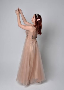 Peri tanrıçası kostümü gibi kremalı fantezi elbisesi ve kristal taç giyen kızıl saçlı bir kızın tam boy portresi. Hafif stüdyo arka planında izole edilmiş zarif hareketli duruş..