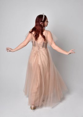 Peri tanrıçası kostümü gibi kremalı fantezi elbisesi ve kristal taç giyen kızıl saçlı bir kızın tam boy portresi. Hafif stüdyo arka planında izole edilmiş zarif hareketli duruş..