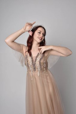 Peri tanrıçası kostümü gibi kremalı fantezi elbisesi ve kristal taç giyen kızıl saçlı bir kızın tam boy portresi. Duruş zarif jest elleri ile, ışık stüdyosunda izole