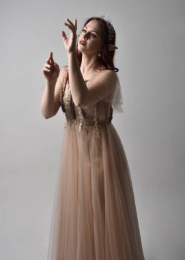 Peri tanrıçası kostümü gibi kremalı fantezi elbisesi ve kristal taç giyen kızıl saçlı bir kızın tam boy portresi. Duruş zarif jest elleri ile, ışık stüdyosunda izole