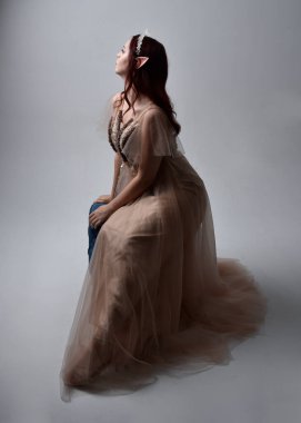Full length  portrait of red haired  girl wearing a creamy fantasy gown and crystal crown, like a fairy goddess costume.  sitting pose with elegant gestural hands, isolated on light studio background. clipart