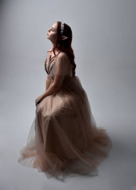 Peri tanrıçası kostümü gibi kremalı fantezi elbisesi ve kristal taç giyen kızıl saçlı bir kızın tam boy portresi. Oturma pozu zarif hareketli eller, hafif stüdyo arka planında izole..