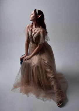 Full length  portrait of red haired  girl wearing a creamy fantasy gown and crystal crown, like a fairy goddess costume.  sitting pose with elegant gestural hands, isolated on light studio background. clipart