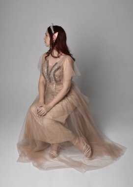 Peri tanrıçası kostümü gibi kremalı fantezi elbisesi ve kristal taç giyen kızıl saçlı bir kızın tam boy portresi. Oturma pozu zarif hareketli eller, hafif stüdyo arka planında izole..