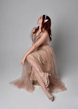 Peri tanrıçası kostümü gibi kremalı fantezi elbisesi ve kristal taç giyen kızıl saçlı bir kızın tam boy portresi. Oturma pozu zarif hareketli eller, hafif stüdyo arka planında izole..