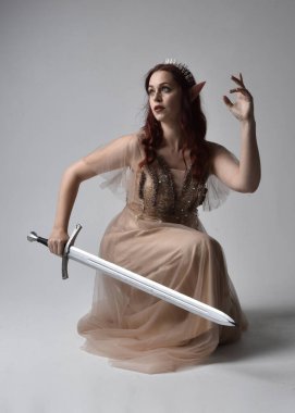 Peri tanrıçası kostümü gibi kremalı fantezi elbisesi ve kristal taç giyen kızıl saçlı bir kızın tam boy portresi. Işık stüdyosunun arka planında izole edilmiş bir kılıçla oturuyor..