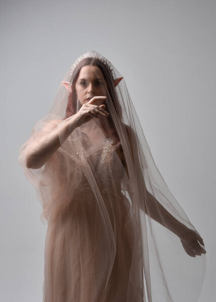 Портрет рыжеволосой девушки в сливочном фэнтези-халате и текстуре вуали, как костюм феи-богини. Стоя поза с элегантной жесты руки, изолированные на светлом фоне студии.