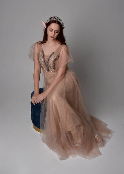 Полноразмерный портрет рыжеволосой девушки в кремовом фантазийном платье и хрустальной короне, как костюм богини-феи. сидя поза с элегантными жестикулярными руками, изолированные на светлом фоне студии.