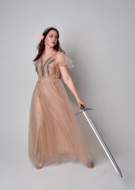Peri tanrıçası kostümü gibi kremalı fantezi elbisesi ve kristal taç giyen kızıl saçlı bir kızın tam boy portresi. Stüdyonun arka planında izole edilmiş bir kılıçla dikiliyordu..