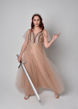 Peri tanrıçası kostümü gibi kremalı fantezi elbisesi ve kristal taç giyen kızıl saçlı bir kızın tam boy portresi. Stüdyonun arka planında izole edilmiş bir kılıçla dikiliyordu..