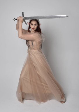 Peri tanrıçası kostümü gibi kremalı fantezi elbisesi ve kristal taç giyen kızıl saçlı bir kızın tam boy portresi. Stüdyonun arka planında izole edilmiş bir kılıçla dikiliyordu..