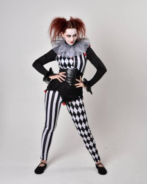 Siyah beyaz palyaço kostümü giyen kızıl saçlı bir kızın uzun bir portresi. Tiyatral sirk karakteri. Stüdyo arka planında izole edilmiş poz.