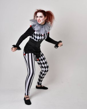 Siyah beyaz palyaço kostümü giyen kızıl saçlı bir kızın uzun bir portresi. Tiyatral sirk karakteri. Stüdyo arka planında izole edilmiş poz.