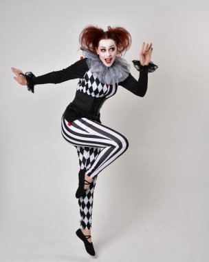 Siyah beyaz palyaço kostümü giyen kızıl saçlı bir kızın uzun bir portresi. Tiyatral sirk karakteri. Stüdyo arka planında izole edilmiş poz.