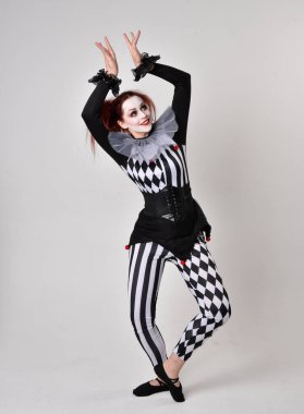 Siyah beyaz palyaço kostümü giyen kızıl saçlı bir kızın uzun bir portresi. Tiyatral sirk karakteri. Stüdyo arka planında izole edilmiş poz.