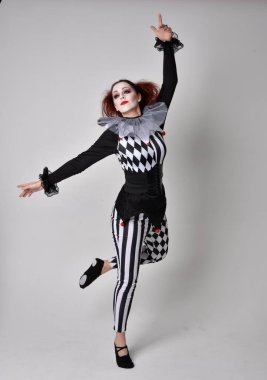 Siyah beyaz palyaço kostümü giyen kızıl saçlı bir kızın uzun bir portresi. Tiyatral sirk karakteri. Stüdyo arka planında izole edilmiş poz.