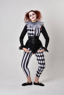 Siyah beyaz palyaço kostümü giyen kızıl saçlı bir kızın uzun bir portresi. Tiyatral sirk karakteri. Stüdyo arka planında izole edilmiş poz.