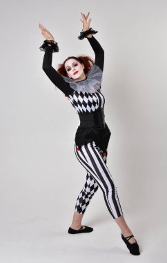 Siyah beyaz palyaço kostümü giyen kızıl saçlı bir kızın uzun bir portresi. Tiyatral sirk karakteri. Stüdyo arka planında izole edilmiş poz.