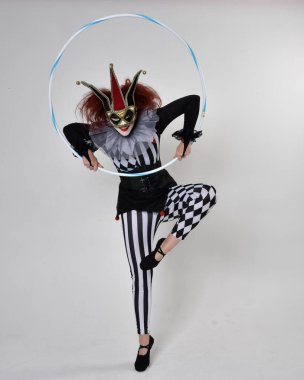 Siyah beyaz palyaço kostümü giyen kızıl saçlı bir kızın uzun bir portresi. Tiyatral sirk karakteri. Stüdyo arka planında izole edilmiş, hula hoop ile poz vermek.