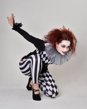 Siyah beyaz palyaço kostümü giyen kızıl saçlı bir kızın uzun bir portresi. Tiyatral sirk karakteri. Stüdyo arka planında izole bir şekilde yerde oturuyor..