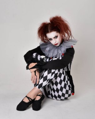 Siyah beyaz palyaço kostümü giyen kızıl saçlı bir kızın uzun bir portresi. Tiyatral sirk karakteri. Stüdyo arka planında izole bir şekilde yerde oturuyor..