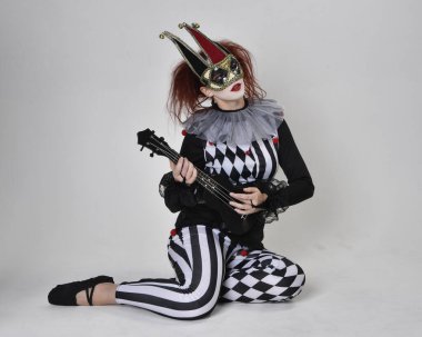 Siyah beyaz palyaço kostümü giyen kızıl saçlı bir kızın uzun bir portresi. Tiyatral sirk karakteri. Stüdyo arka planında izole bir şekilde yerde oturuyor..