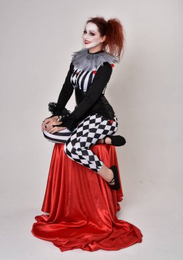 Siyah beyaz palyaço kostümü giyen kızıl saçlı bir kızın uzun bir portresi. Tiyatral sirk karakteri. Oturma pozu, stüdyo arka planında izole.