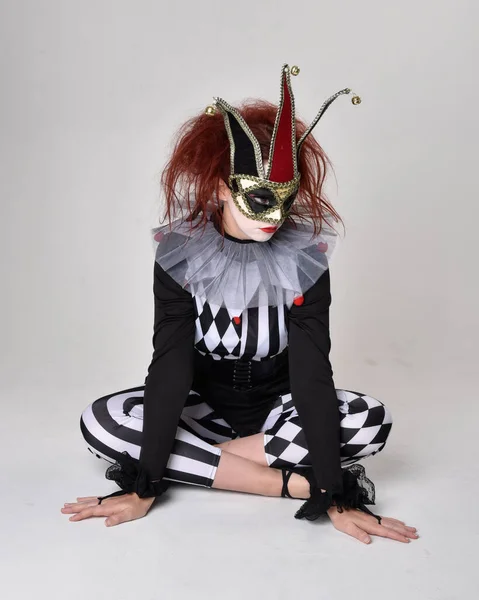 全长的红头发女孩肖像穿着黑白相间的小丑小丑戏服 戏剧化的马戏团角色 在地板上坐下 隔离在工作室的背景下 — 图库照片