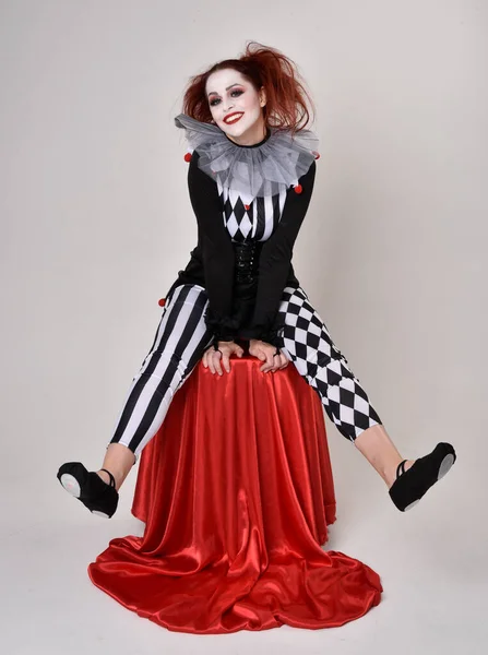 黒と白のピエロジェスターの衣装 演劇サーカスのキャラクターを身に着けている赤い髪の少女の完全な長さの肖像画 座位姿勢はスタジオ背景に隔離され — ストック写真