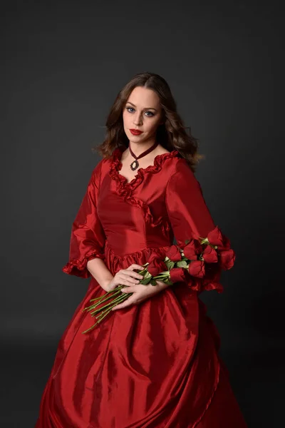 portrait of pretty female model with red hair wearing glamorous historical victorian red ballgown.  Posing with a moody dark background.