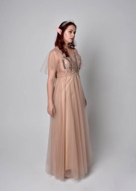 Kızıl saçlı, göz kamaştırıcı fantezi tül cübbe ve taç giyen güzel bir kadın modelin tam boy portresi. Stüdyo arka planında hareketli kollarla poz vermek