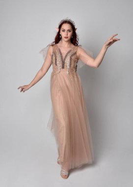 Kızıl saçlı, göz kamaştırıcı fantezi tül cübbe ve taç giyen güzel bir kadın modelin tam boy portresi. Stüdyo arka planında hareketli kollarla poz vermek