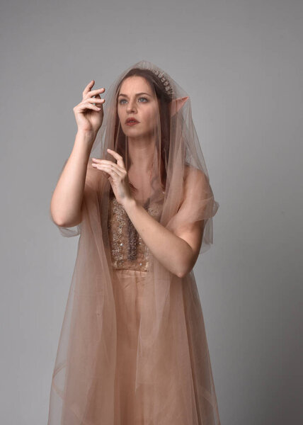  Полноразмерный портрет красивой женщины-модели с рыжими волосами в гламурном фэнтезийном тюлевом платье, короне и плащанице. Пэддок с жестикуляционными руками на студийном фоне