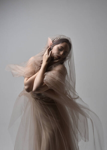  Full length portrait of pretty female model with red hair wearing glamorous fantasy tulle gown, crown and shroud veil.  Posing with gestural arms on a studio background