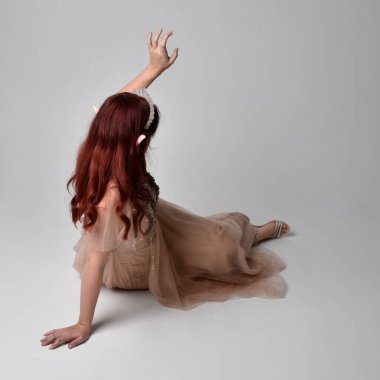 Kızıl saçlı, çekici fantezi tül cübbe, taç ve kefen peçeli güzel bir kadın modelin tam boy portresi. Stüdyonun arka planında diz çöküp poz vermek